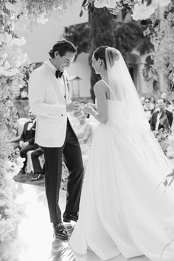 Ring exchange as bride and groom face each other under a floral arch, guests seated behind in an outdoor courtyard with trees