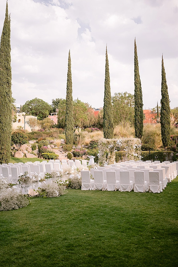Ceremony setup for an outdoor wedding ceremony with semi circle chair seating, white aisle runner, and floral arch on a lawn by cypress trees