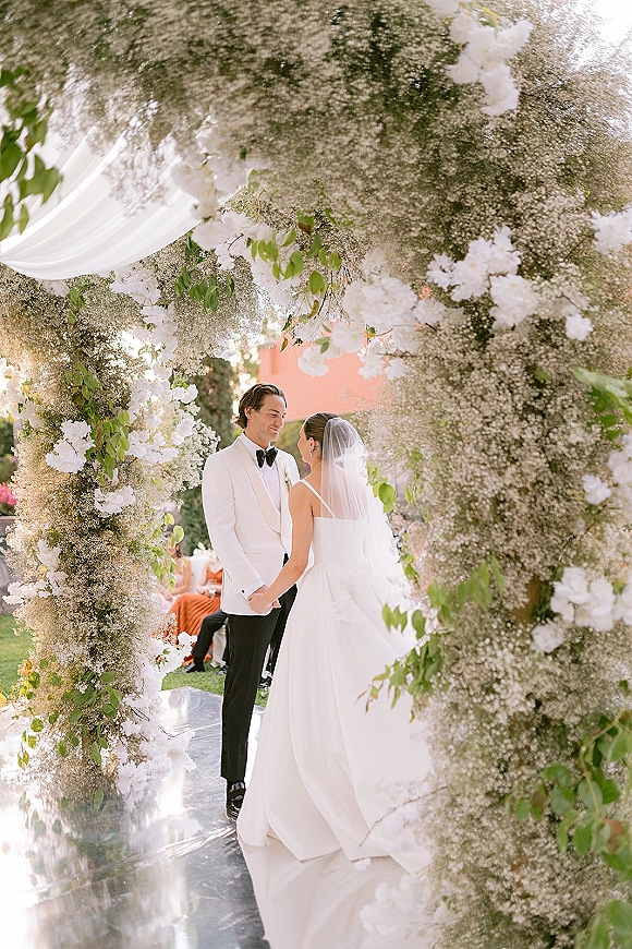 Ceremony moment as couple holding hands during wedding vows beneath a white floral arch with draped fabric in an outdoor garden