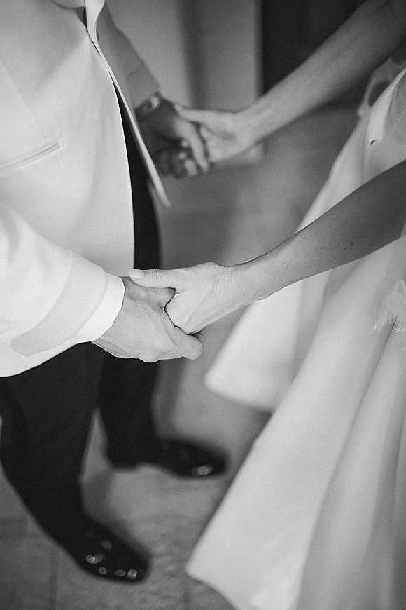Wedding handholding as bride and groom clasp hands, showing dress fabric and groom suit, tie and wristwatch near an indoor doorway