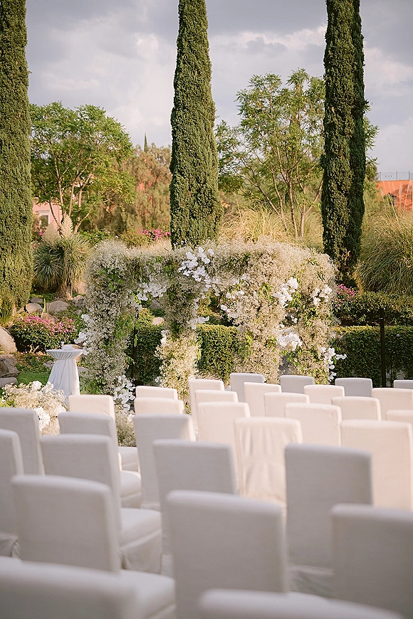 Ceremony setup for an outdoor wedding ceremony with a white floral arch, hanging florals and greenery, and white aisle chairs in a hedge garden with cypress trees