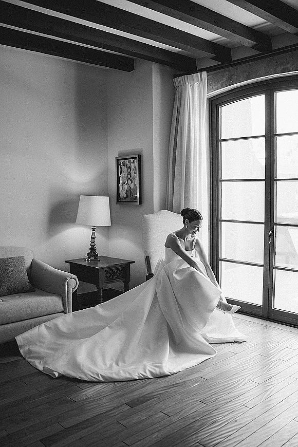 Bridal portrait in black and white of a bride putting on high heels, long train draped as she sits by a large window in a chair