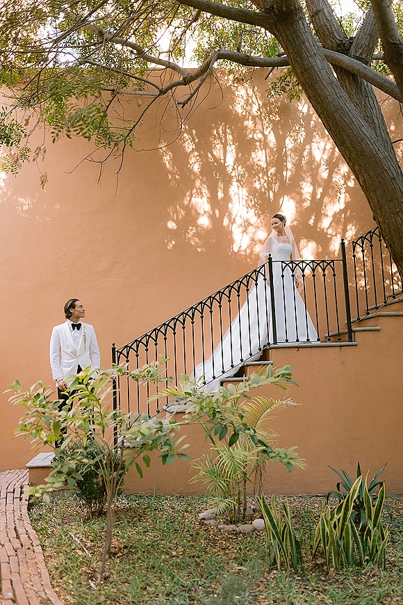 Couple portrait of groom in white tuxedo looking up at bride on staircase, her long train flowing beside iron railing by stucco wall