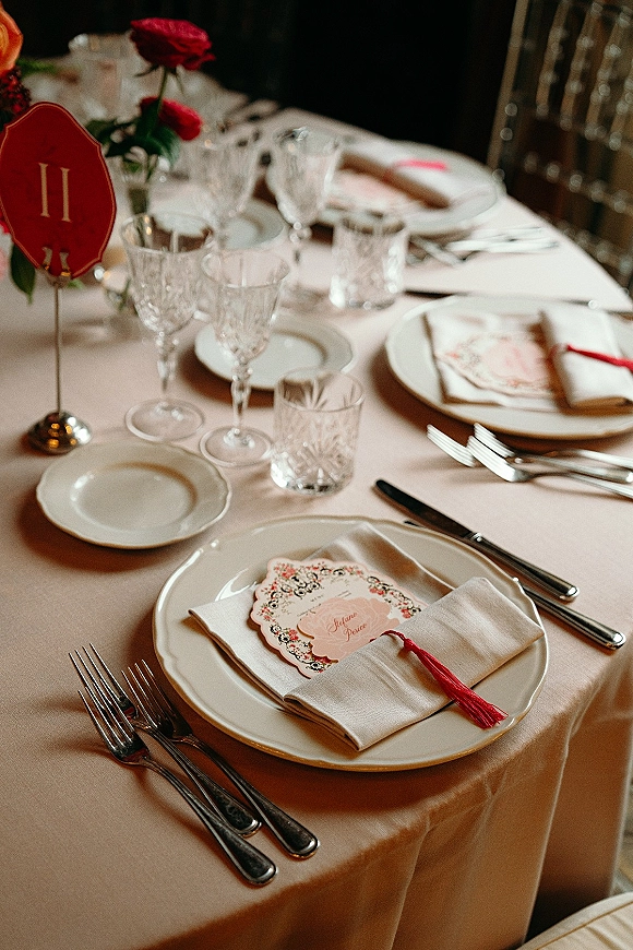 Reception tablescape with blush wedding table decor featuring white plates, crystal goblets, silver flatware, and red rose bud vase indoors