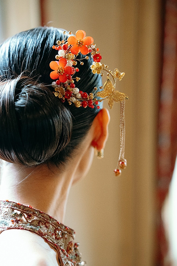 Bridal hair accessory with a wedding hairpiece of gold flowers, pearls, and red beads draped over a sleek low bun in warm indoor light