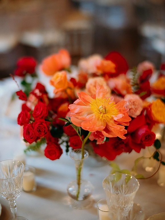 Wedding centerpiece with red rose centerpiece and coral blooms in a bud vase, surrounded by votive candles and water goblets on a white tablecloth