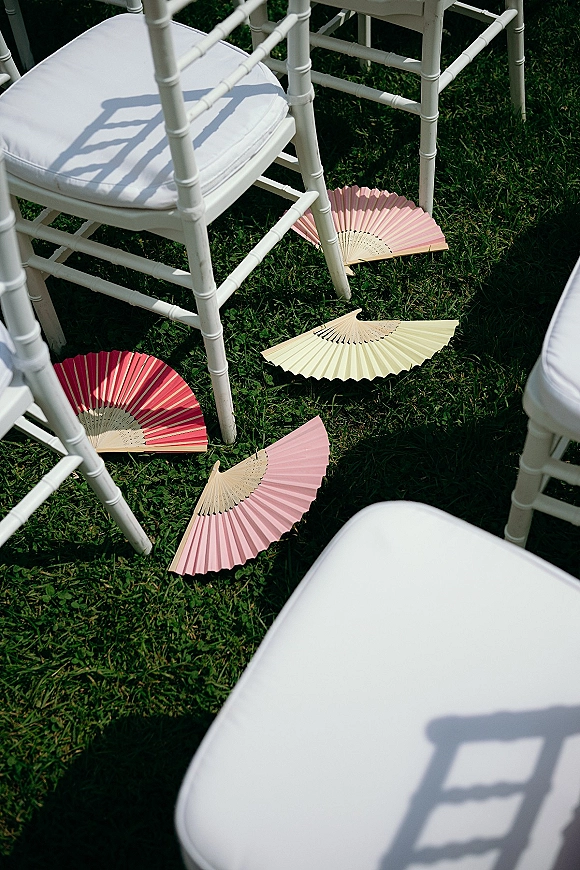 Ceremony fan favors on white Chiavari chairs with chair cushions, paper folding fans neatly set for guests on a grass lawn
