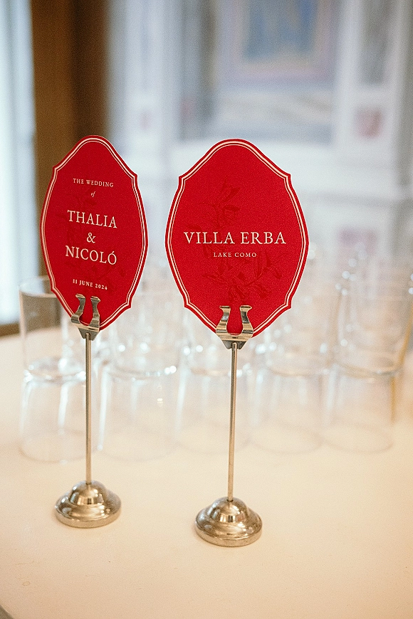 Wedding place cards in red with calligraphy on silver holders beside table number cards on a white-clothed reception table near windows