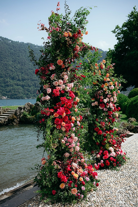 Wedding floral installation with roses and greenery forming an asymmetrical ceremony floral backdrop by a lakeside with mountain views under blue sky