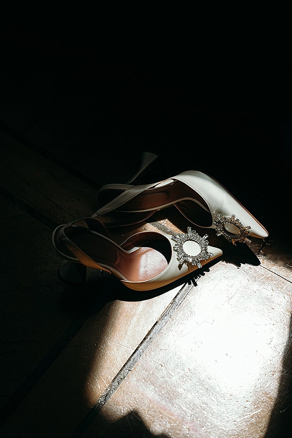 Bridal shoes in ivory satin slingback heels with crystal shoe clips on a wood floor, sunlit shadows in a dark interior