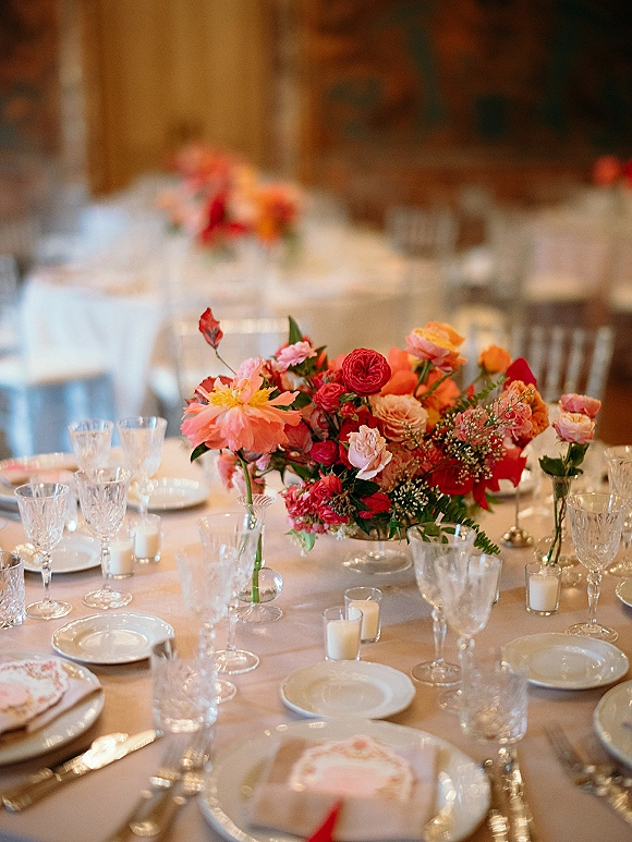 Reception tablescape with a wedding table centerpiece of coral and pink blooms, bud vases, and votive candles on round tables in a warm banquet hall