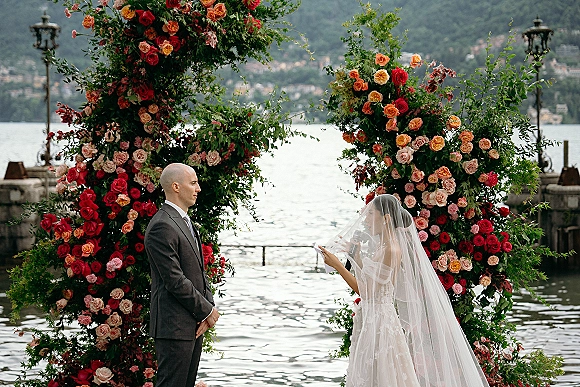 Wedding vows as bride reading vows to groom beneath a rose and greenery arch on a stone terrace overlooking a mountain lake