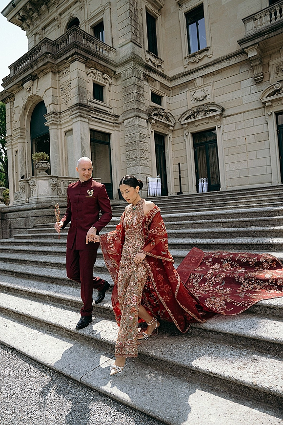 Couple portrait of bride and groom holding hands, descending stone steps by an ornate historic facade, her red embroidered robe trailing long train
