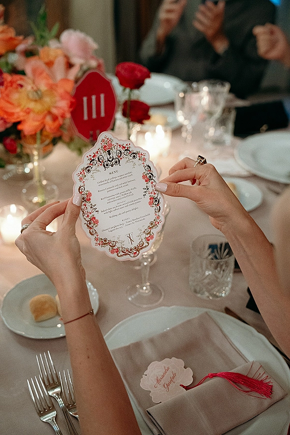 Wedding menu card with scalloped edges and floral border held over a candlelit reception tablescape with red tassel place card and centerpiece