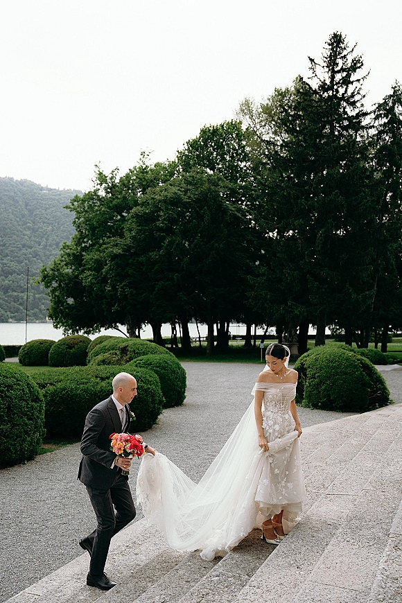Couple portrait of bride and groom walking up stone steps, groom holding veil, bride with pink bouquet and long train in garden setting