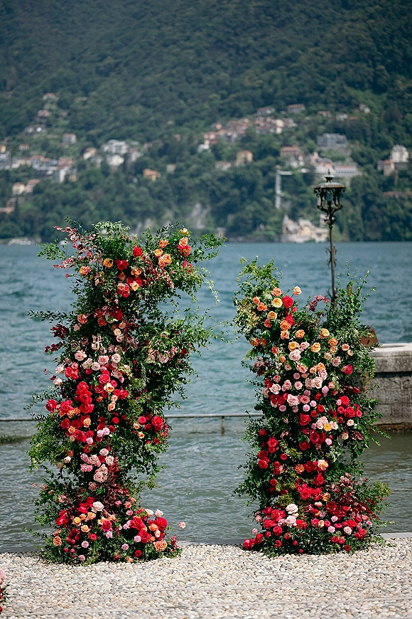 Wedding ceremony arch with floral ceremony pillars of red and pink roses and lush greenery on a stone terrace overlooking a lake and hills