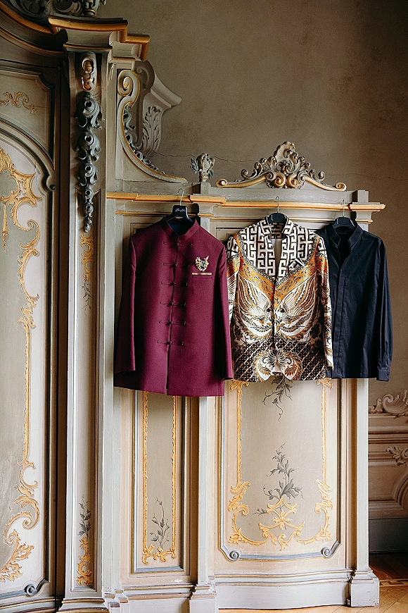 Groom attire flat lay with burgundy sherwani jacket, patterned and black jackets on hangers, floral boutonniere on ornate vintage wardrobe backdrop
