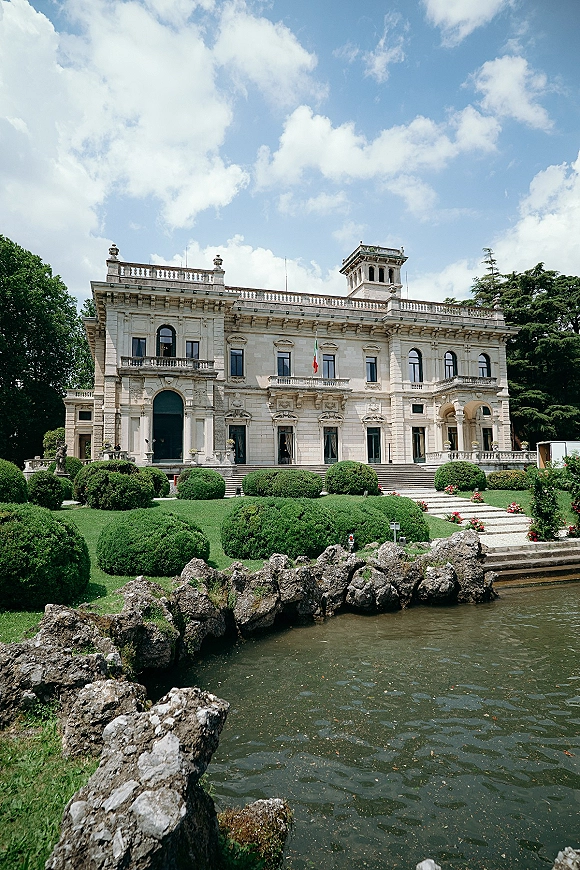 Wedding venue exterior with stone balustrades and arched doorways, framed by manicured hedges and a pond under a blue sky