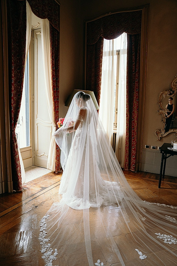Bridal portrait of a bride by window holding a bouquet, her long cathedral veil trailing on hardwood floors in soft window light