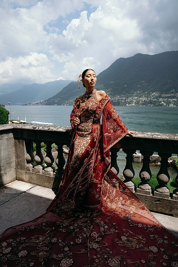 Bridal portrait of a bride in a red wedding dress with gold-embroidered long train and sheer cape on a stone balcony above a lake and mountains
