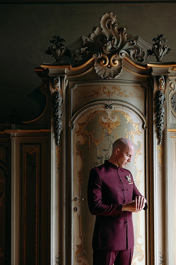 Groom portrait with groom adjusting watch in a burgundy suit and boutonniere, posed by an ornate gilded armoire in a vintage room.