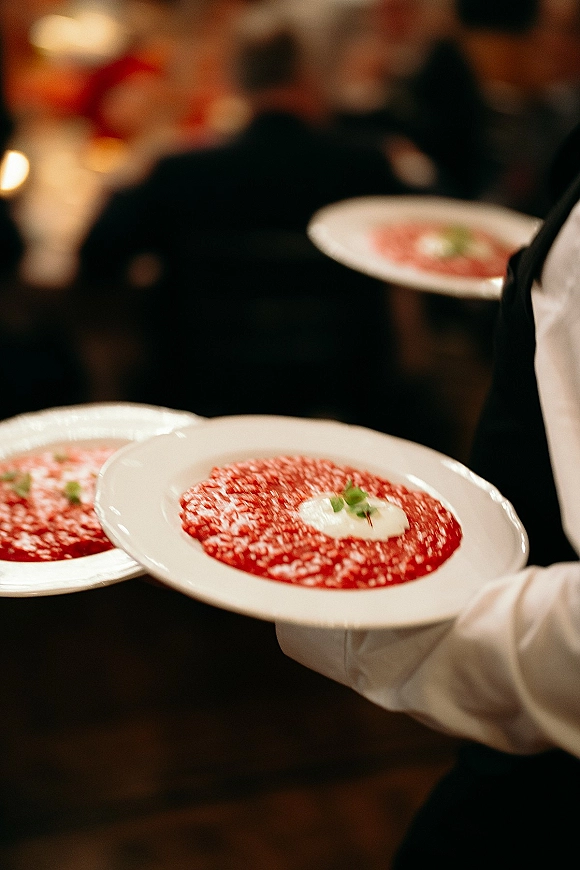 Wedding plated dinner served by a uniformed server carrying white plates of red soup with cream and herb garnish in a dim banquet room