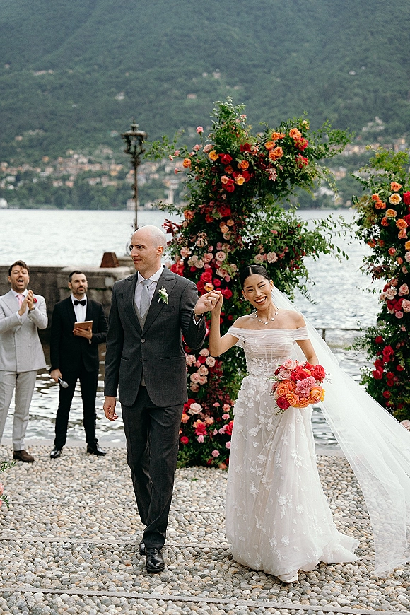 Wedding recessional as bride and groom walk hand in hand past a rose floral arch on a lakeside stone terrace with mountains behind