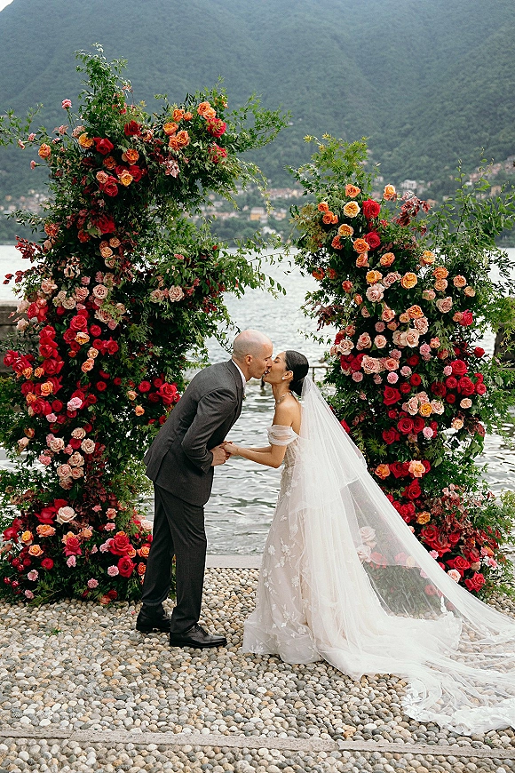 Wedding kiss portrait of bride and groom kissing beneath a floral arch, veil flowing, with lake and mountains behind on pebbled shore