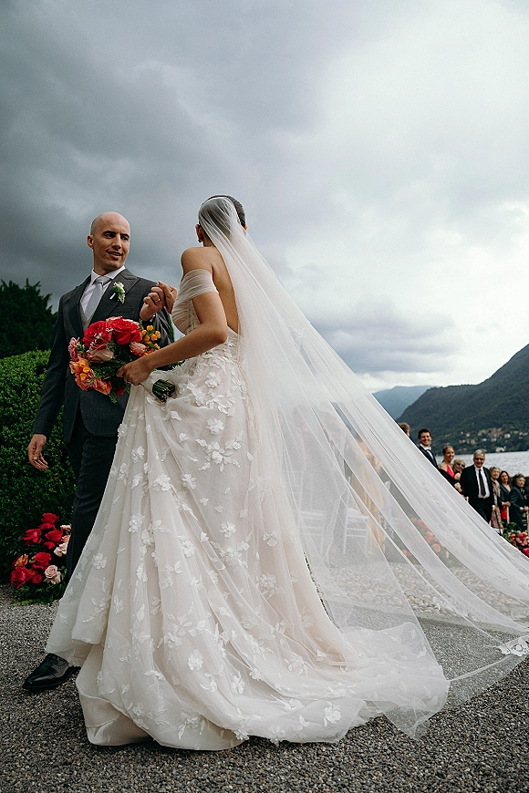 Ceremony moment as bride and groom walking aisle, her cathedral veil and lace dress trailing toward a mountain lake under cloudy sky