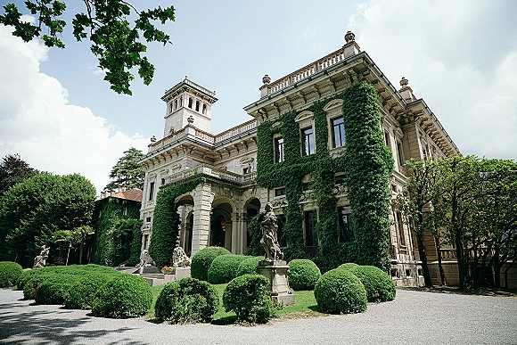 Wedding venue exterior with an ivy covered facade, arched entryway, stone statues, and a gravel driveway framed by manicured hedges under clouds