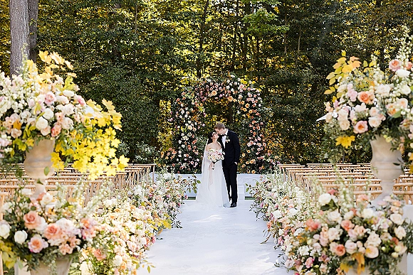 Ceremony moment as bride and groom kiss under a floral arch with a blush accent, sunlit forest garden aisle and white runner