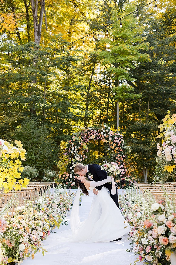 Wedding kiss as groom dips bride in a strapless dress holding bouquet beneath a floral arch with string lights in sunlit forest ceremony aisle