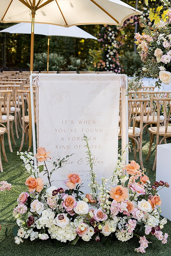 Wedding ceremony signage with a fabric welcome sign and pastel roses and hydrangeas, set on a garden lawn by wooden chairs and umbrellas