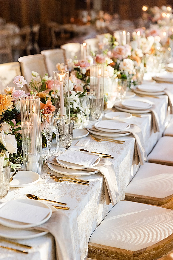 Reception tablescape with long head table decor featuring floral garland, taper candles, crystal glassware, gold flatware in warm candlelit room