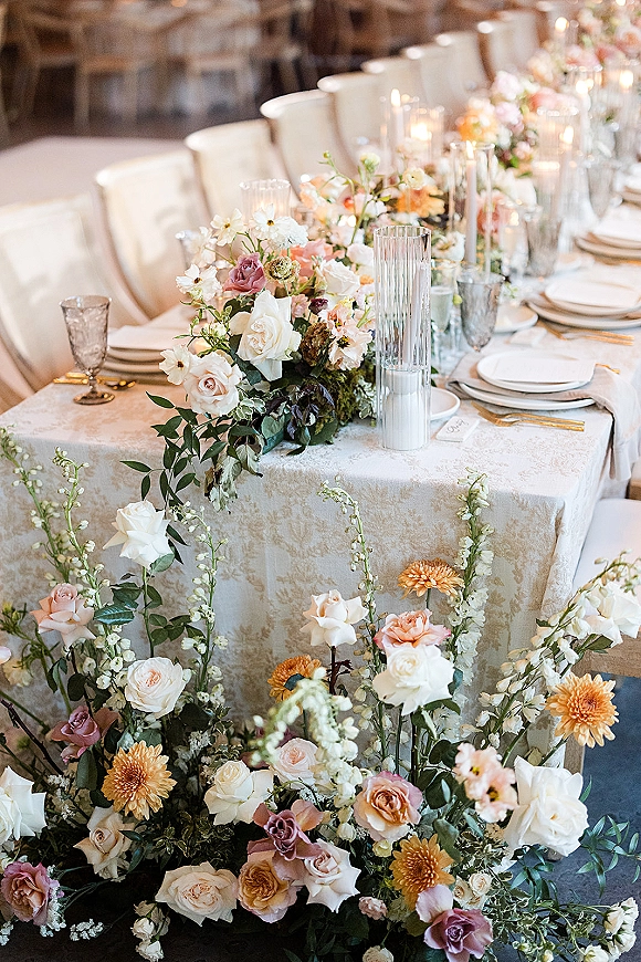 Reception tablescape with wedding head table decor featuring a floral runner, taper candles, white plates, gold flatware, and goblets indoors