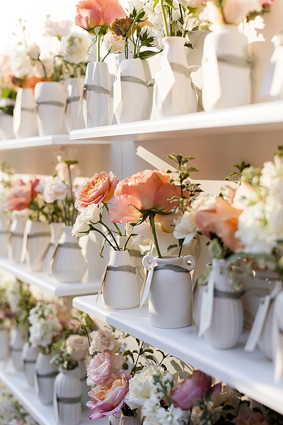 Wedding floral display in white ceramic vases with peach roses, white blooms, and greenery on white shelves in a sunlit outdoor setting