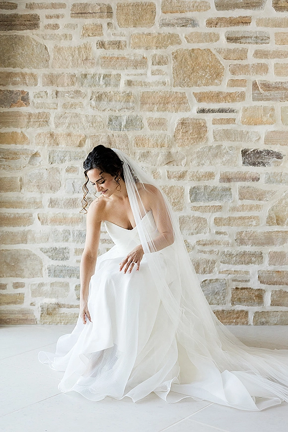 Bridal portrait of a bride in a strapless wedding dress with a long veil, seated on a white floor against a stone wall