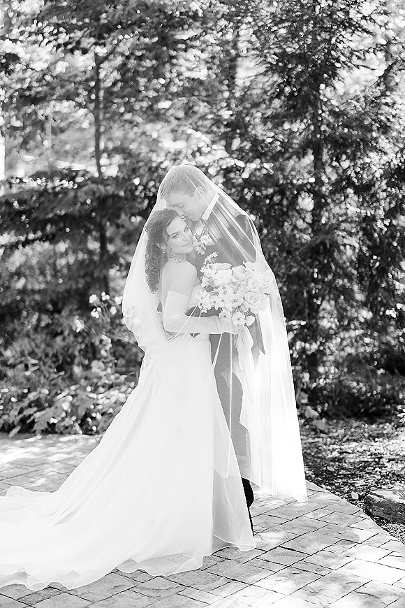 Couple portrait, black and white wedding portrait of groom kissing bride’s forehead under veil as she holds bouquet on a garden stone path