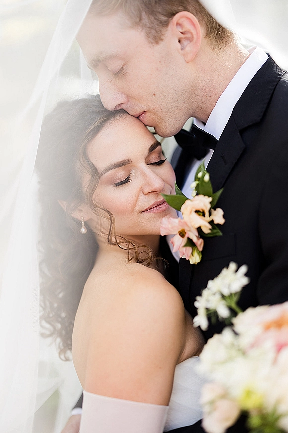 Wedding couple portrait of groom kissing bride’s forehead as she stands under a bridal veil, holding a bouquet against a soft neutral backdrop