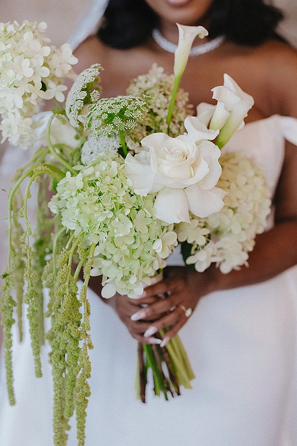 Bridal bouquet of white hydrangea and calla lilies with roses and trailing amaranthus, held by bride in gown with pearl necklace