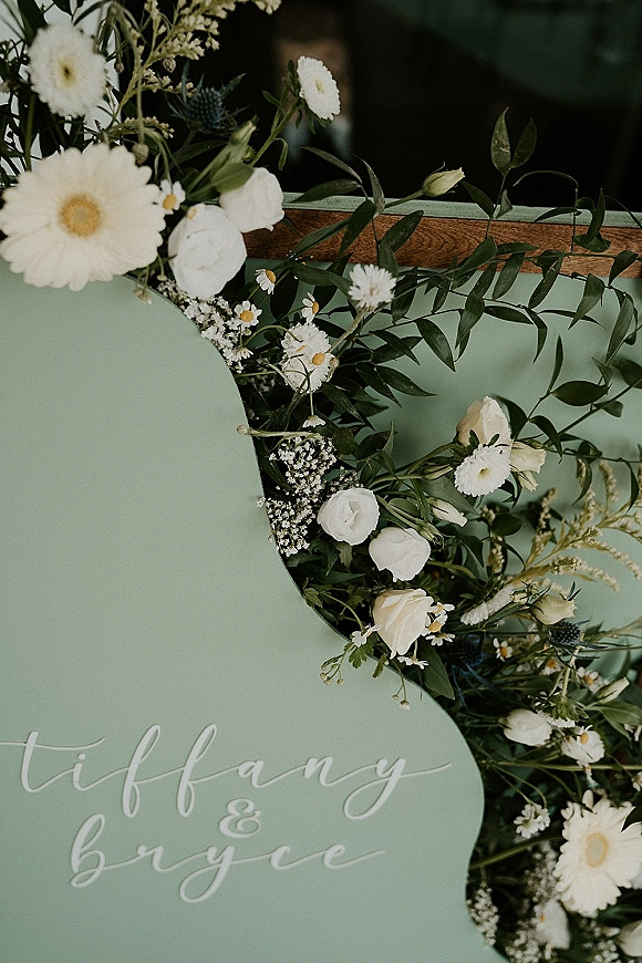 Wedding welcome sign with green wedding welcome sign lettering, framed by white daisies, baby's breath, and eucalyptus garland against a dark indoor background