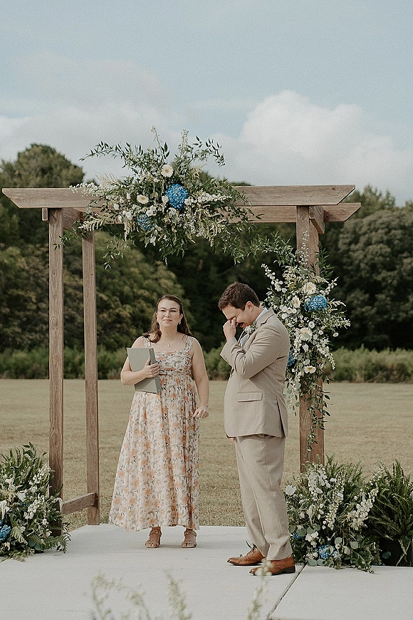 Ceremony moment as groom in a tan suit wipes tears beside the officiant under a wood arch with blue hydrangea accents on a lawn
