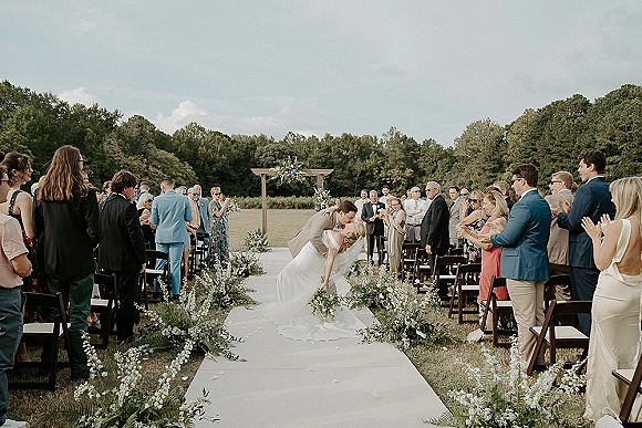 Ceremony kiss at an outdoor wedding ceremony as the bride and groom kiss beneath a wood arch with greenery, guests standing in a field