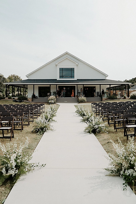 Ceremony aisle design with a white aisle runner lined by ground florals and greenery, leading to a white barn porch under string lights