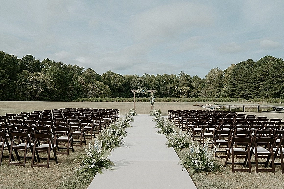 Ceremony setup for an outdoor wedding ceremony with a white aisle runner, floral-lined chairs, and a wooden arbor by a pond and dock