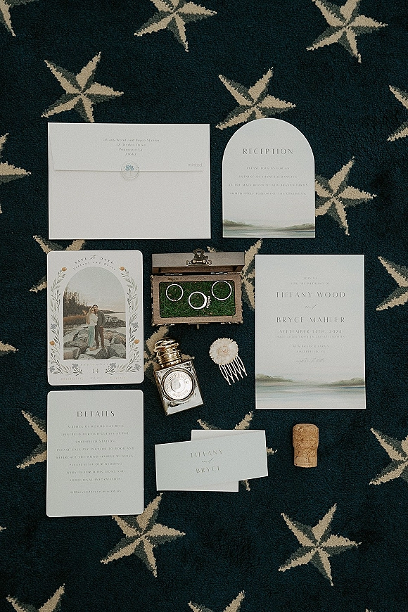 Wedding invitation suite flat lay with envelopes, arched cards, wax seal, wood ring box and perfume on a patterned carpet background