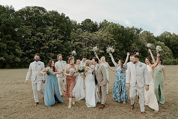 Wedding party photo of bride and groom with wedding party walking in an open field, holding bouquets, with trees and sky behind