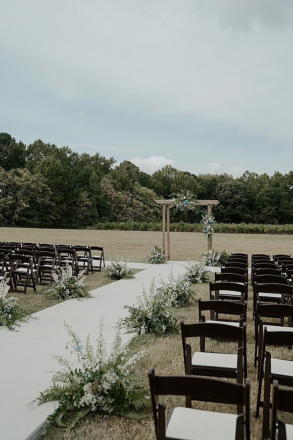 Outdoor ceremony setup with wedding ceremony aisle and wooden wedding arch, white aisle runner and greenery florals in an open field under clouds