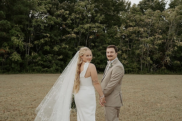 Couple portrait of bride and groom holding hands in a grass field, her long bridal veil and satin dress flowing under an overcast sky