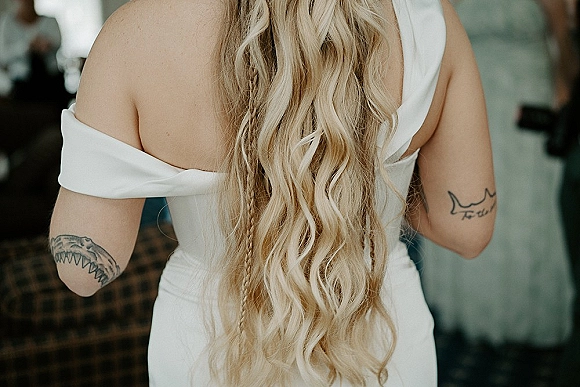 Bridal hairstyle with long wedding hair in loose blonde curls and a braid detail, shown from behind with an off-shoulder gown indoors